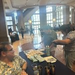 Smiling board member sits next to table with Texas Folklife merchandise. Two people stand in front of the table viewing items.