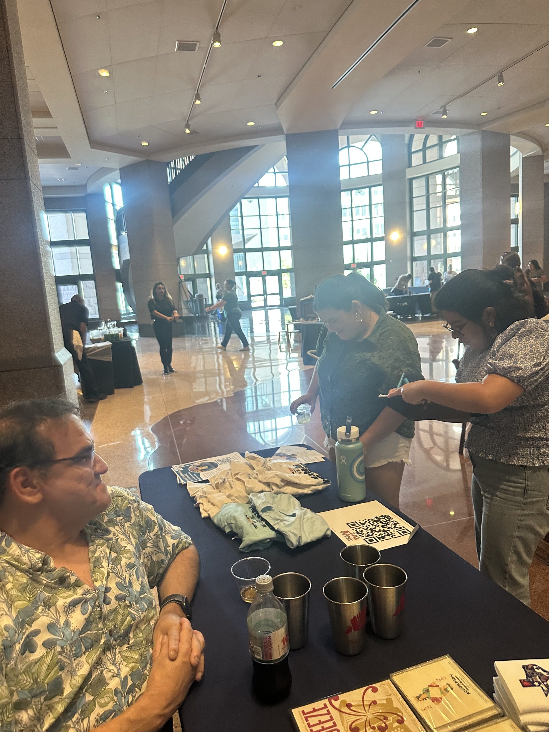 Smiling board member sits next to table with Texas Folklife merchandise. Two people stand in front of the table viewing items.