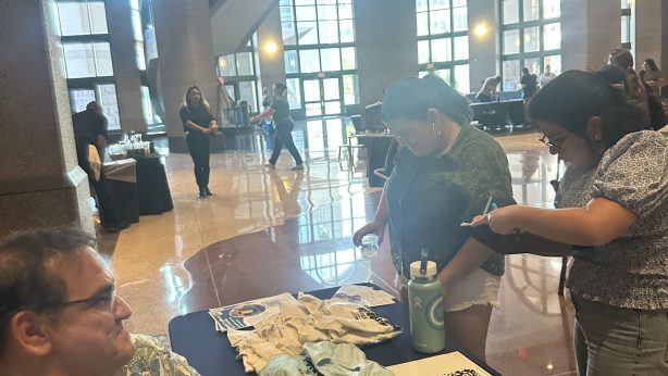 Smiling board member sits next to table with Texas Folklife merchandise. Two people stand in front of the table viewing items.