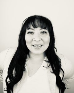 Black and white headshot of Alba Donajhi Sereno, Texas Folklife fellow, smiling at the camera. She has long dark hair with bangs and a small nose stud, and wears a light V-neck top with a light cardigan against a plain background.