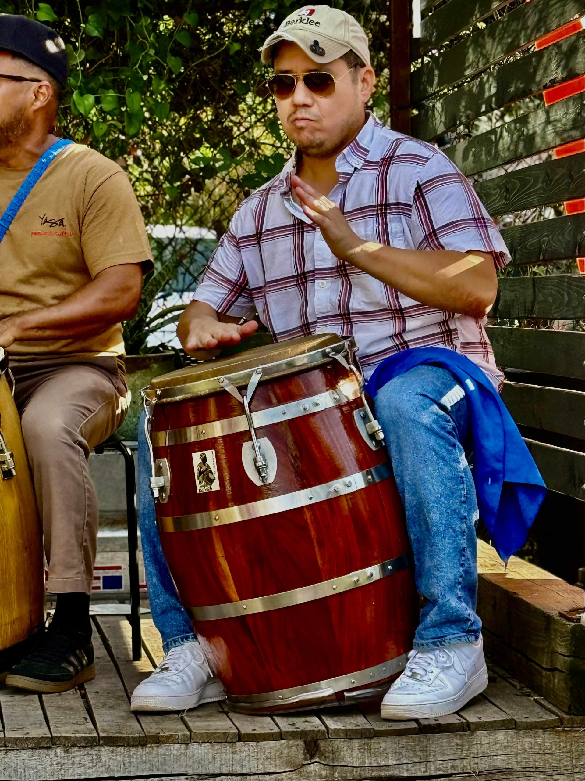 Adam Rosario, wearing a Berklee cap and sunglasses, plays a tall barrel-shaped hand drum outdoors next to another percussionist.