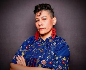 Portrait of Amber Ortega, who has short dark hair styled up on one side and wears large red geometric earrings and a blue floral top. Her arms are crossed and she looks directly at the camera against a gray backdrop.