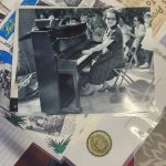 Black and white photograph of a woman in sunglasses and a plaid dress seated at an upright piano outdoors, playing as a group of people sit nearby. The print rests on a layer of aged newspaper clippings.