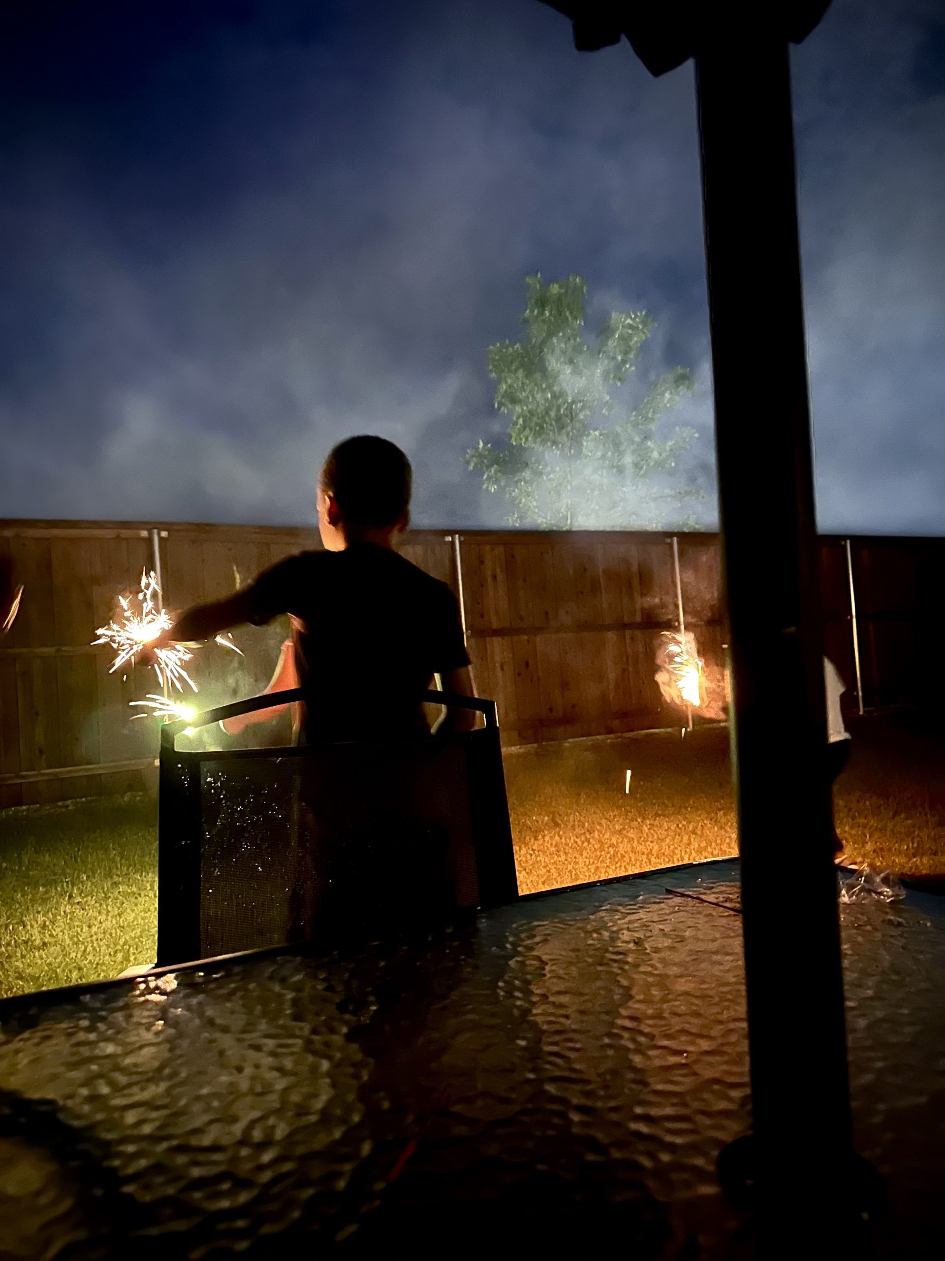 A child holds a glowing sparkler in a backyard at dusk, with a wooden fence behind them.