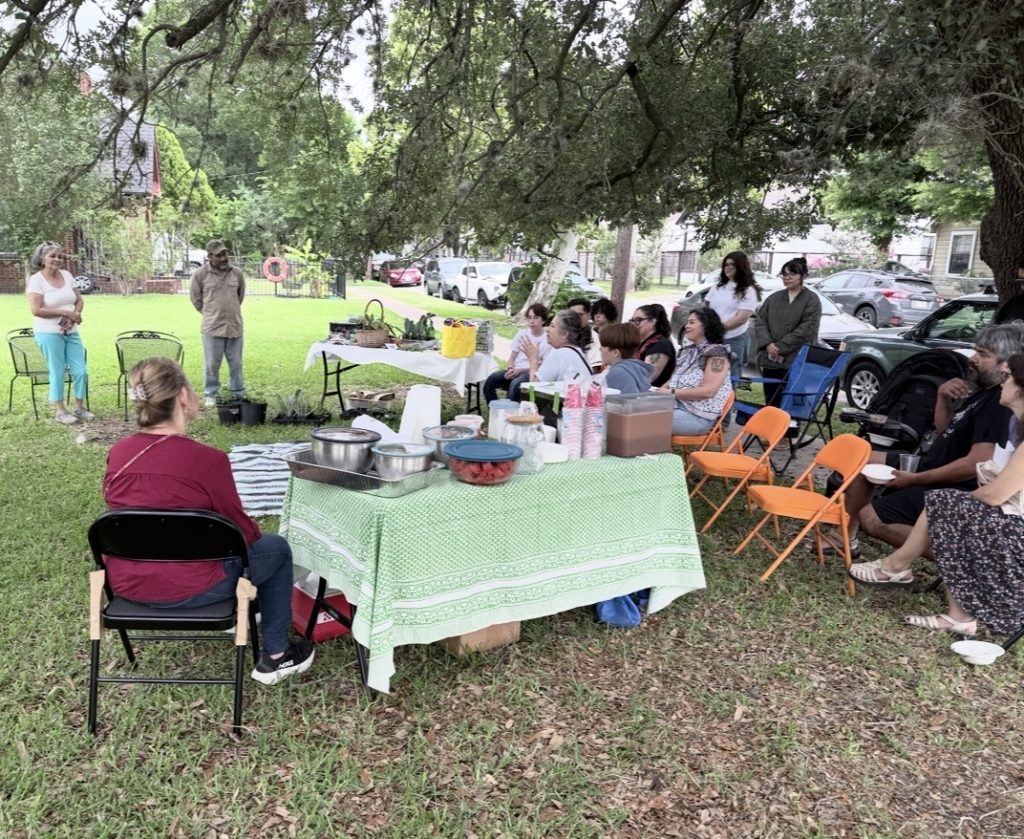 Community members gather in folding chairs beneath a large shade tree for an outdoor seed and plant exchange. A long table holds bowls of food, drinks, and sliced watermelon, with potted seedlings nearby and neighborhood homes in the background.