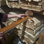 Close-up of stacked wooden slats and boards separated by sheets of aged newspaper on storage shelves. One hand reaches toward the stacks while another holds a long, polished wooden board.