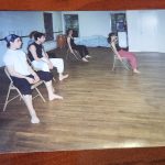 Color photograph of four people seated on metal folding chairs along the wall of a dance studio with wood floors, a ballet barre, and a conga drum, with large windows in the background.
