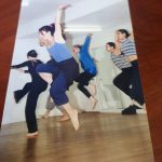 Color photograph of a group of barefoot dancers mid-leap in a studio with wood floors and white walls, arms raised, wearing casual practice clothes.