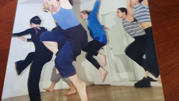 Color photograph of a group of barefoot dancers mid-leap in a studio with wood floors and white walls, arms raised, wearing casual practice clothes.