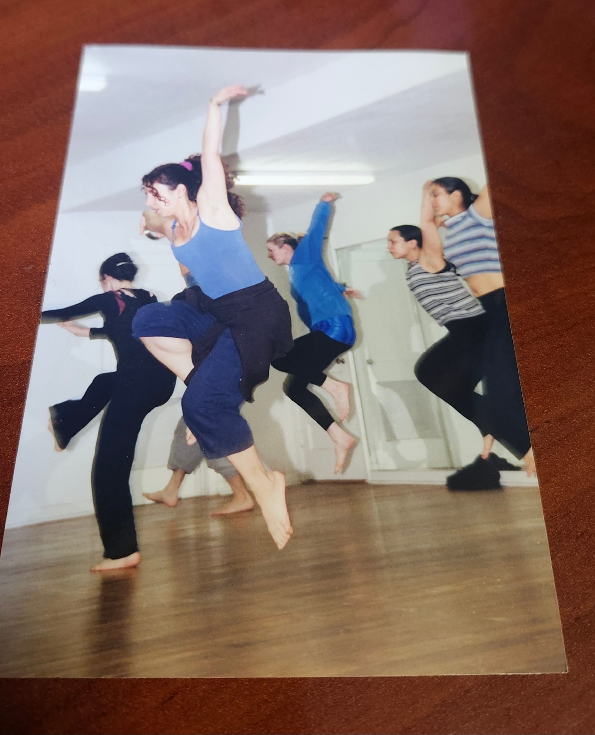 Color photograph of a group of barefoot dancers mid-leap in a studio with wood floors and white walls, arms raised, wearing casual practice clothes.