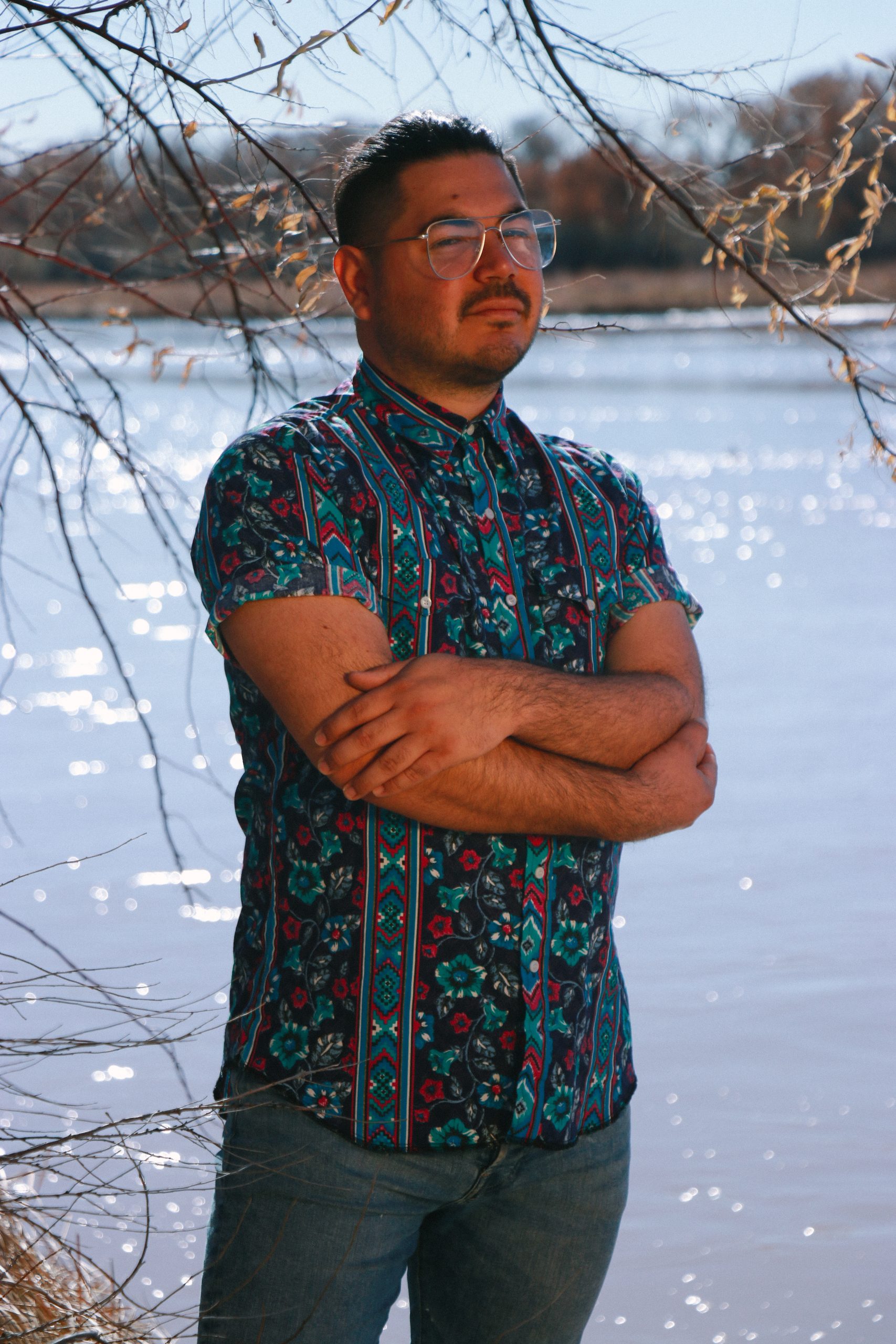Beto De León stands with arms crossed beside a sunlit river, wearing a floral and geometric print shirt and glasses, framed by bare tree branches with autumn trees on the far bank.