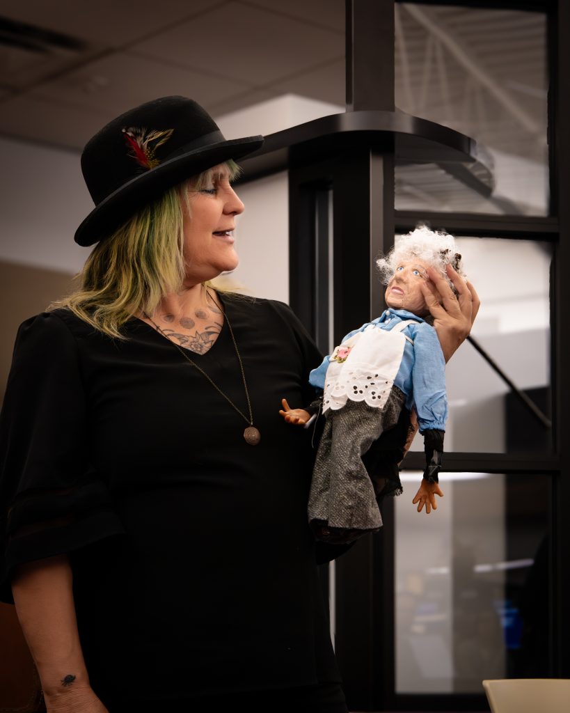 _DSC4701(1) A performer in a black feathered hat holds a hand puppet of an elderly woman with white curly hair, a blue shirt, and a white apron embroidered with a pink flower.