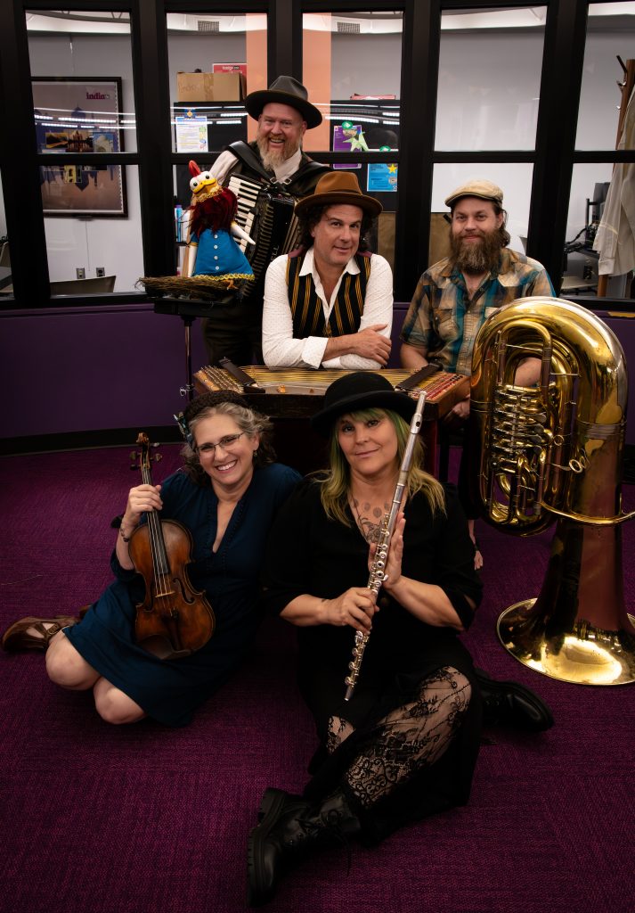 _DSC4828 Five members of the band Mazel Tov Kocktail Hour pose together in a room with tall windows and purple carpet. Three musicians stand behind with an accordion, a hammered dulcimer, and a tuba. Two musicians sit in front holding a violin and a flute. A rooster puppet in a blue dress perches on a stand beside them.