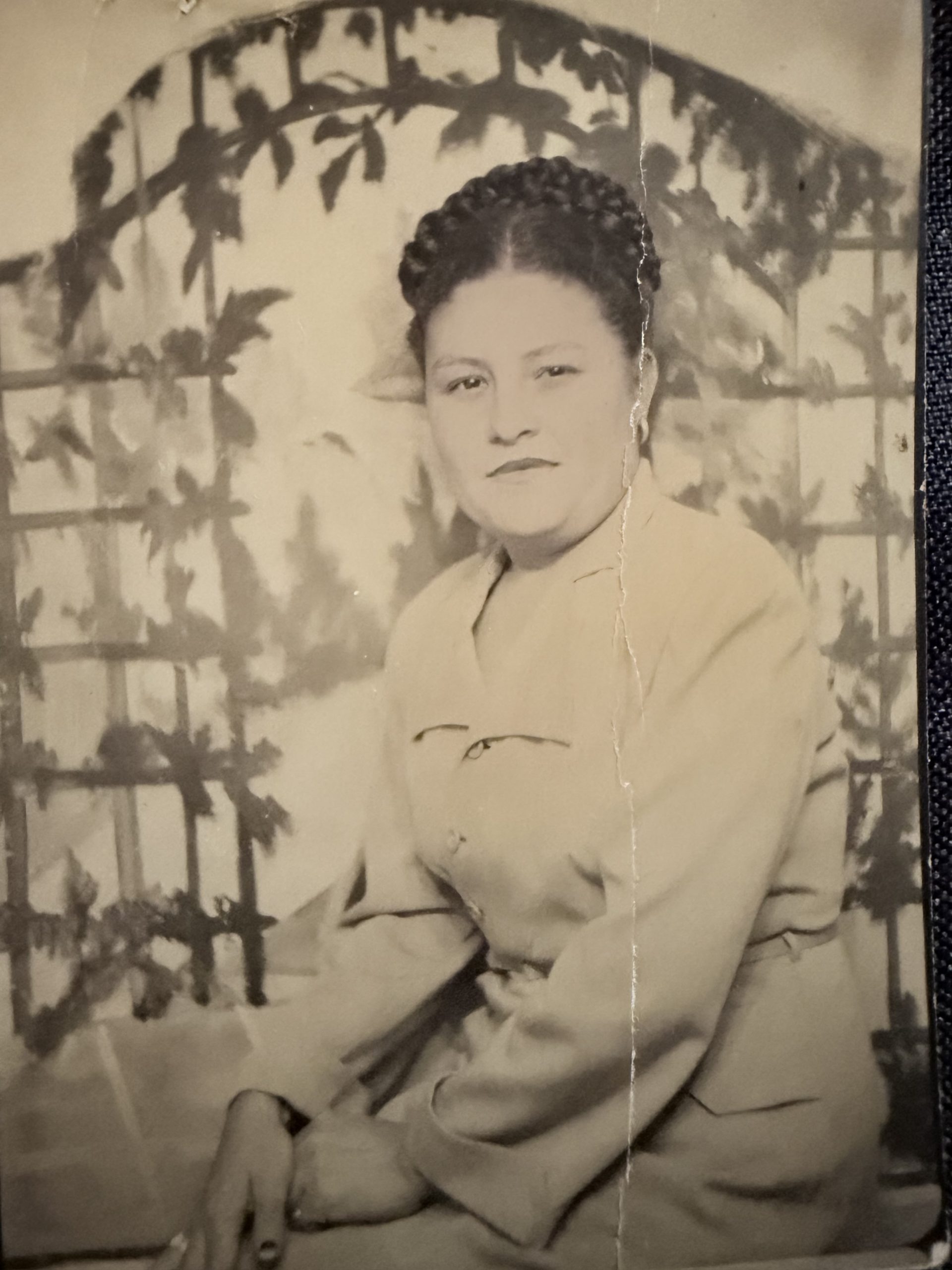 IMG_2151 Sepia-toned vintage portrait of a young woman with her hair in a braided crown, seated in front of a painted garden trellis backdrop. A crease runs down the center of the photograph.