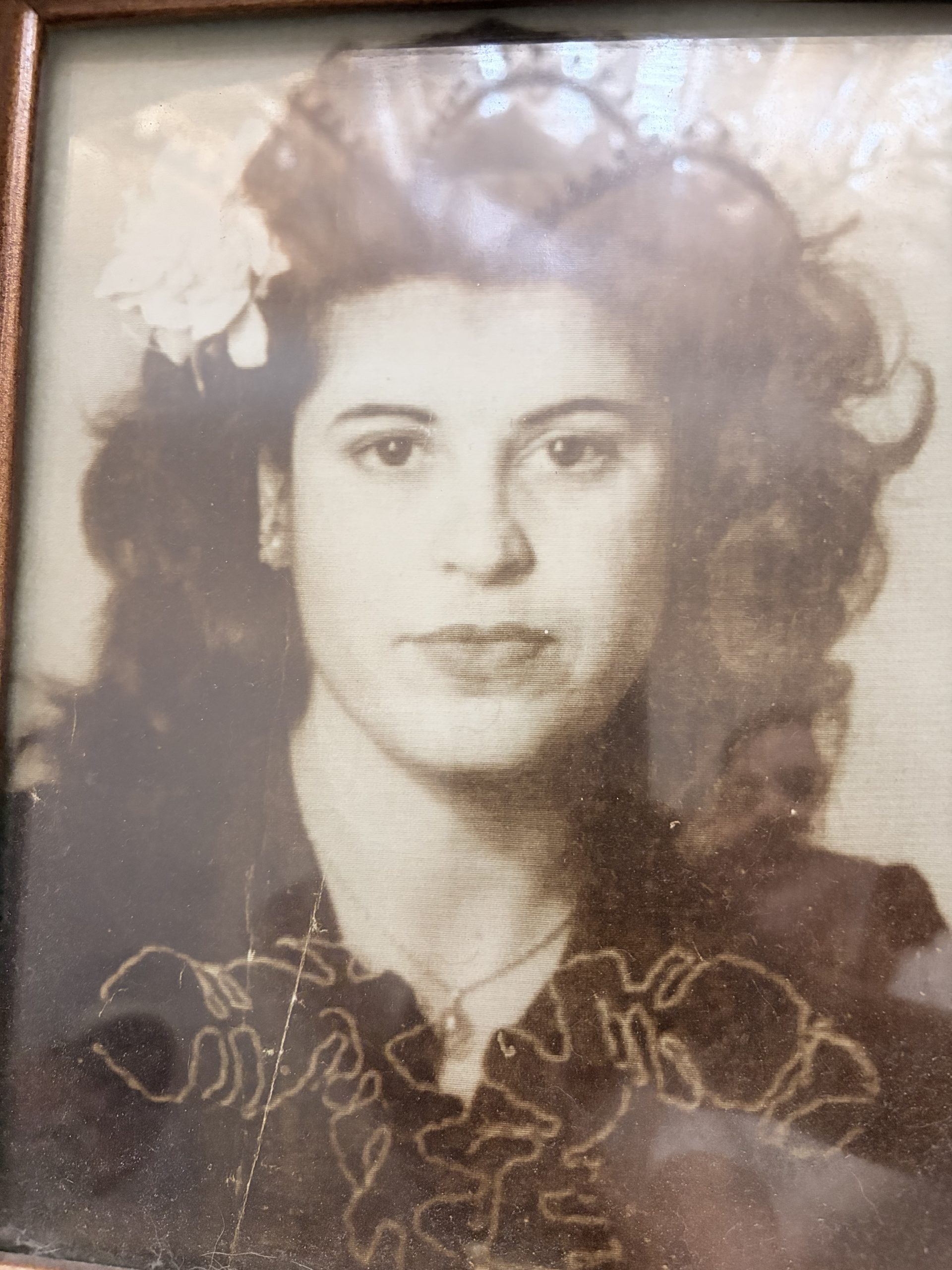 IMG_2152 Framed sepia portrait of a young woman with white flowers pinned in her wavy dark hair, wearing an embroidered dark blouse. The glass reflects light across the image.