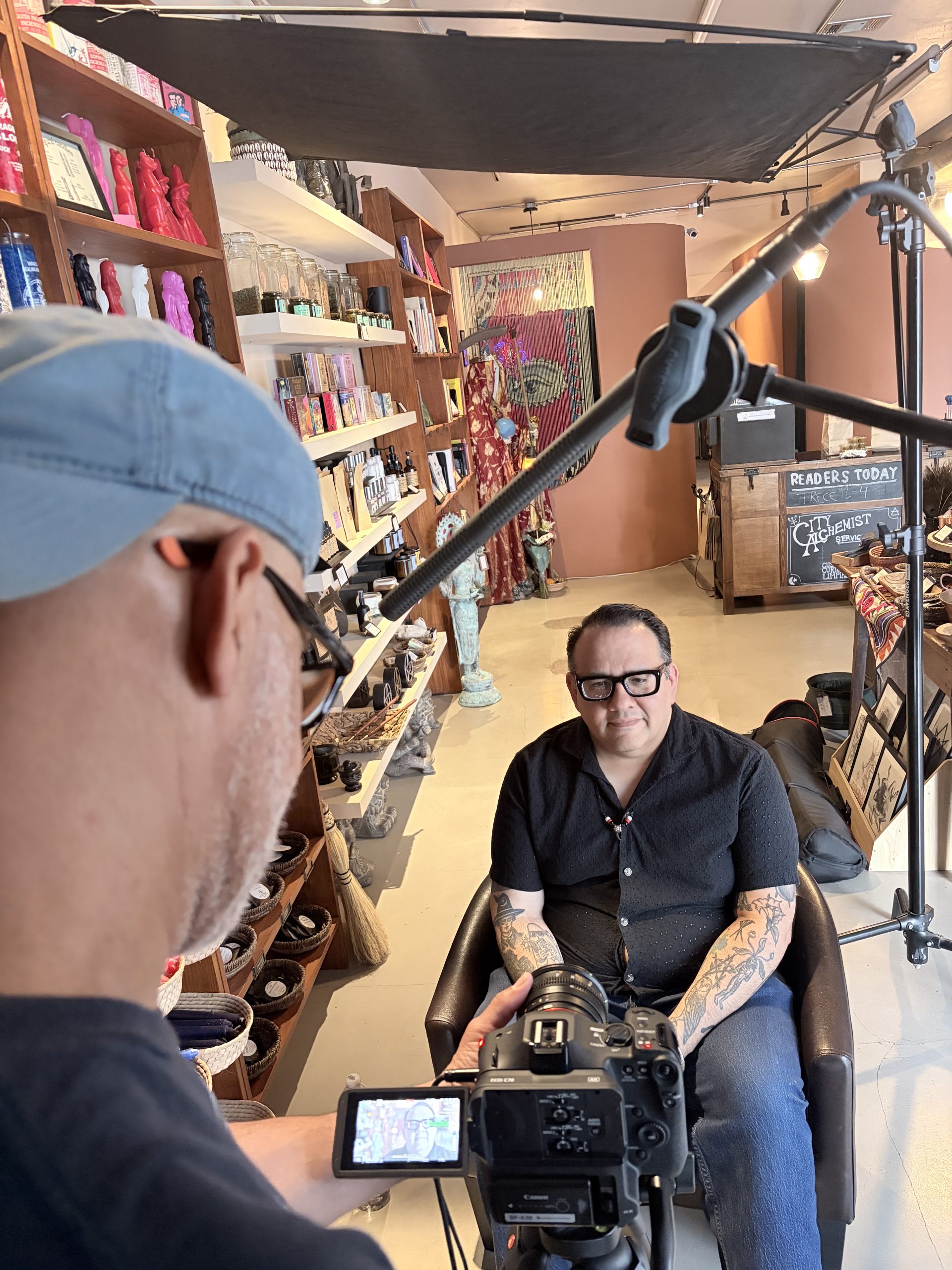 IMG_2167 Alexis Arredondo of City Alchemist sits for an interview inside the shop, surrounded by shelves of ritual candles, herbs, and books. A camera operator films from the foreground, and a chalkboard sign reading "City Alchemist" is visible behind him.