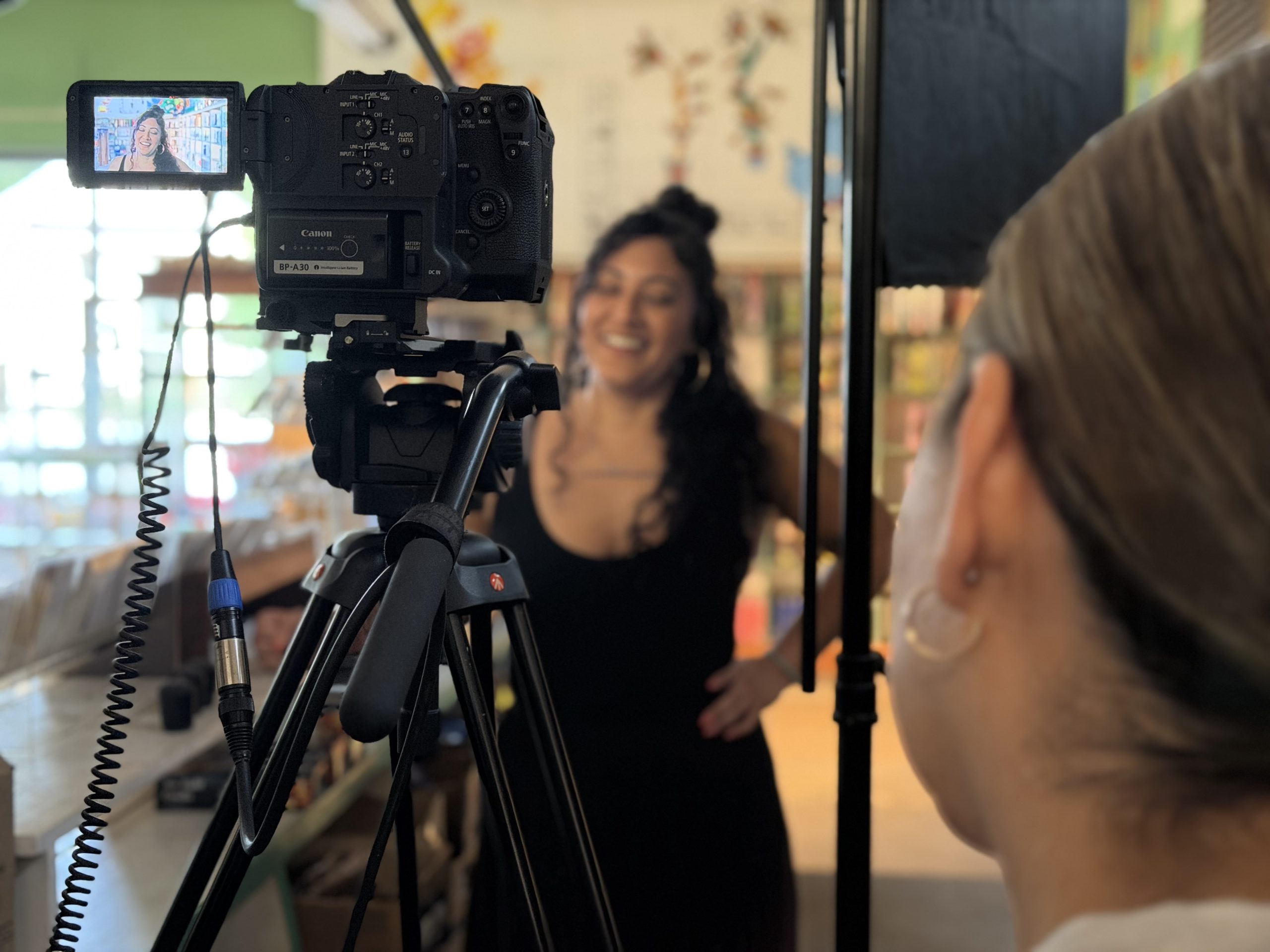 IMG_3355 Elena Gonzales laughs during a filmed interview inside a brightly lit shop. A camera on a tripod frames her in the foreground while a crew member adjusts a light panel nearby.
