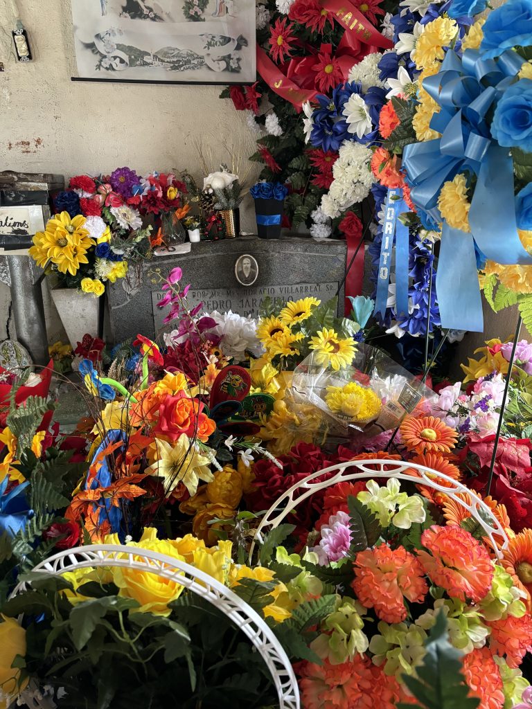 A memorial shrine at Don Pedro Jaramillo's gravesite overflowing with flowers, sunflowers, roses, and ribbons in red, yellow, blue, and orange surrounding a weathered headstone.