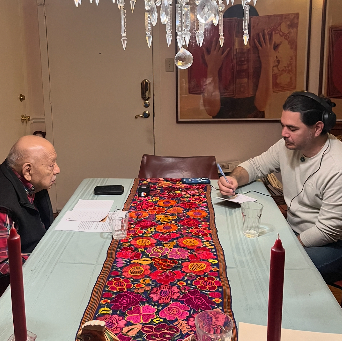 Beto De León interviews Dr. Tomás Ybarra-Frausto at a dining table, headphones on and pen in hand, with a floral embroidered table runner and lit candles between them.