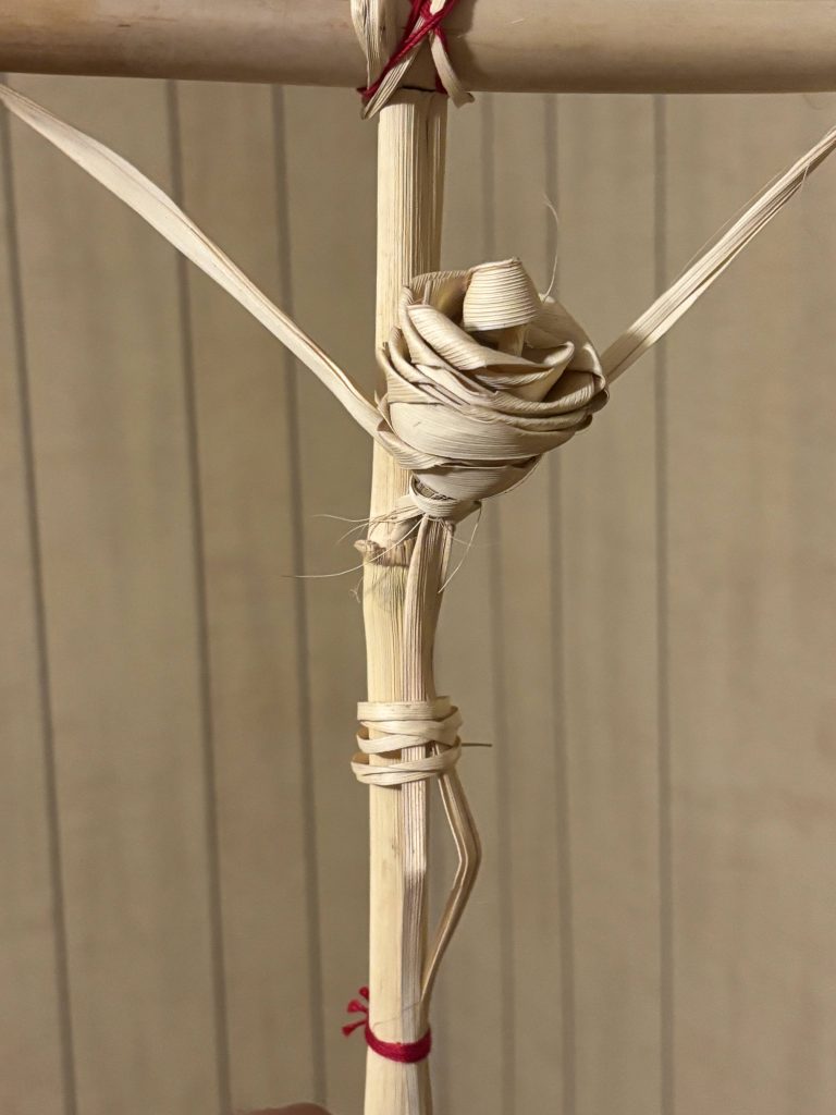 A cross woven from carrizo cane and palm fronds, with a rosette shaped palm flower at its center, tied with red thread.
