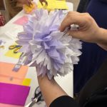 Hands shaping a lavender tissue paper flower at a workshop table scattered with colored paper, scissors, and partly finished blossoms.
