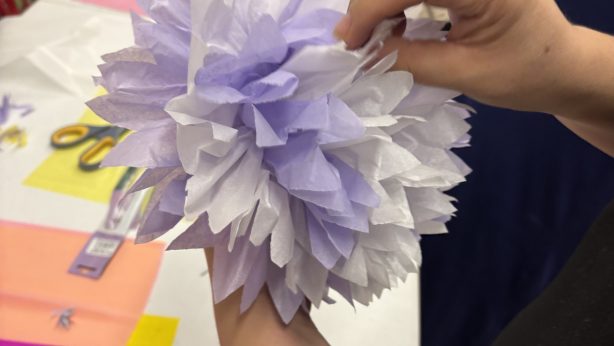 Hands shaping a lavender tissue paper flower at a workshop table scattered with colored paper, scissors, and partly finished blossoms.