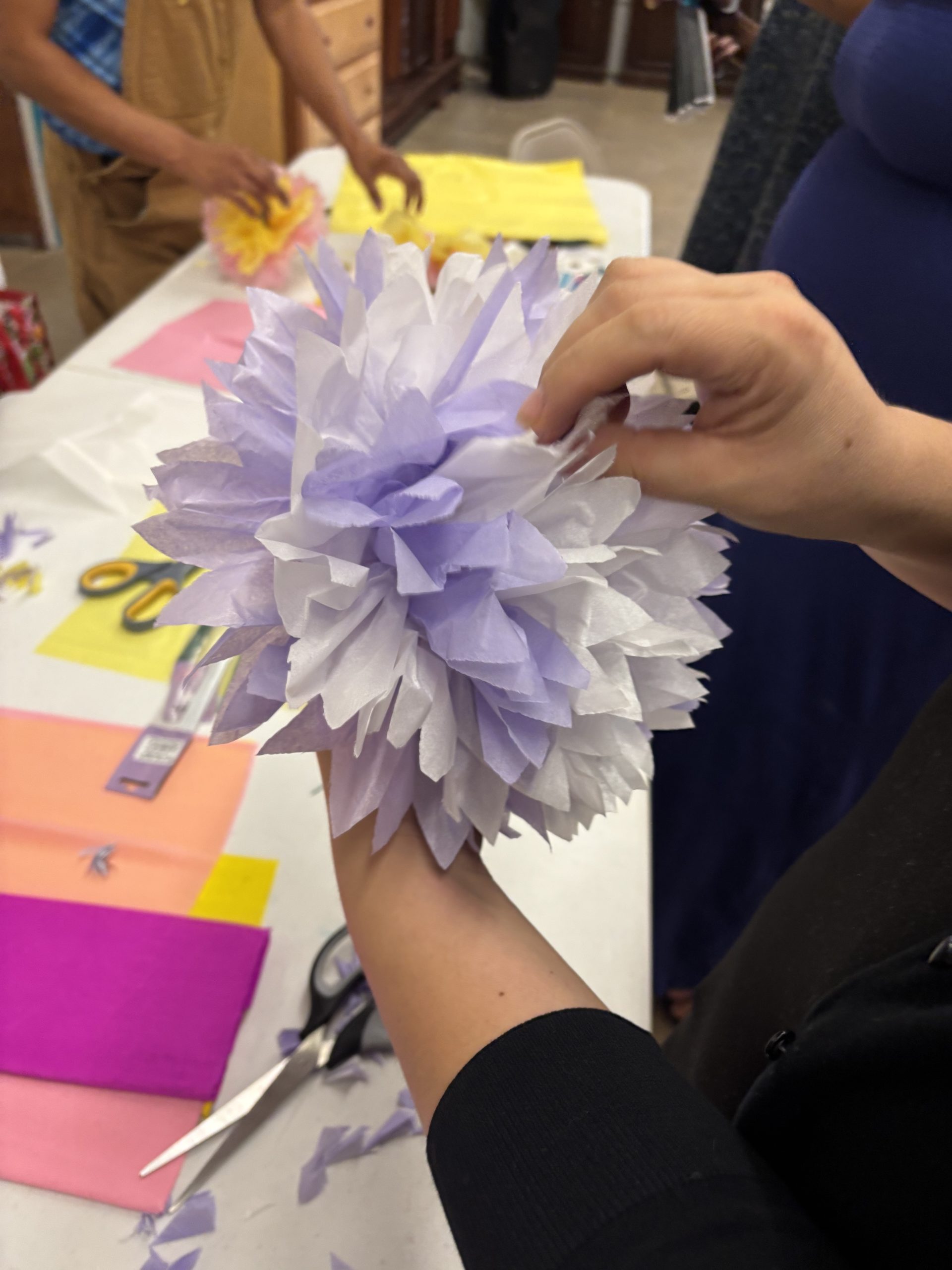 Hands shaping a lavender tissue paper flower at a workshop table scattered with colored paper, scissors, and partly finished blossoms.