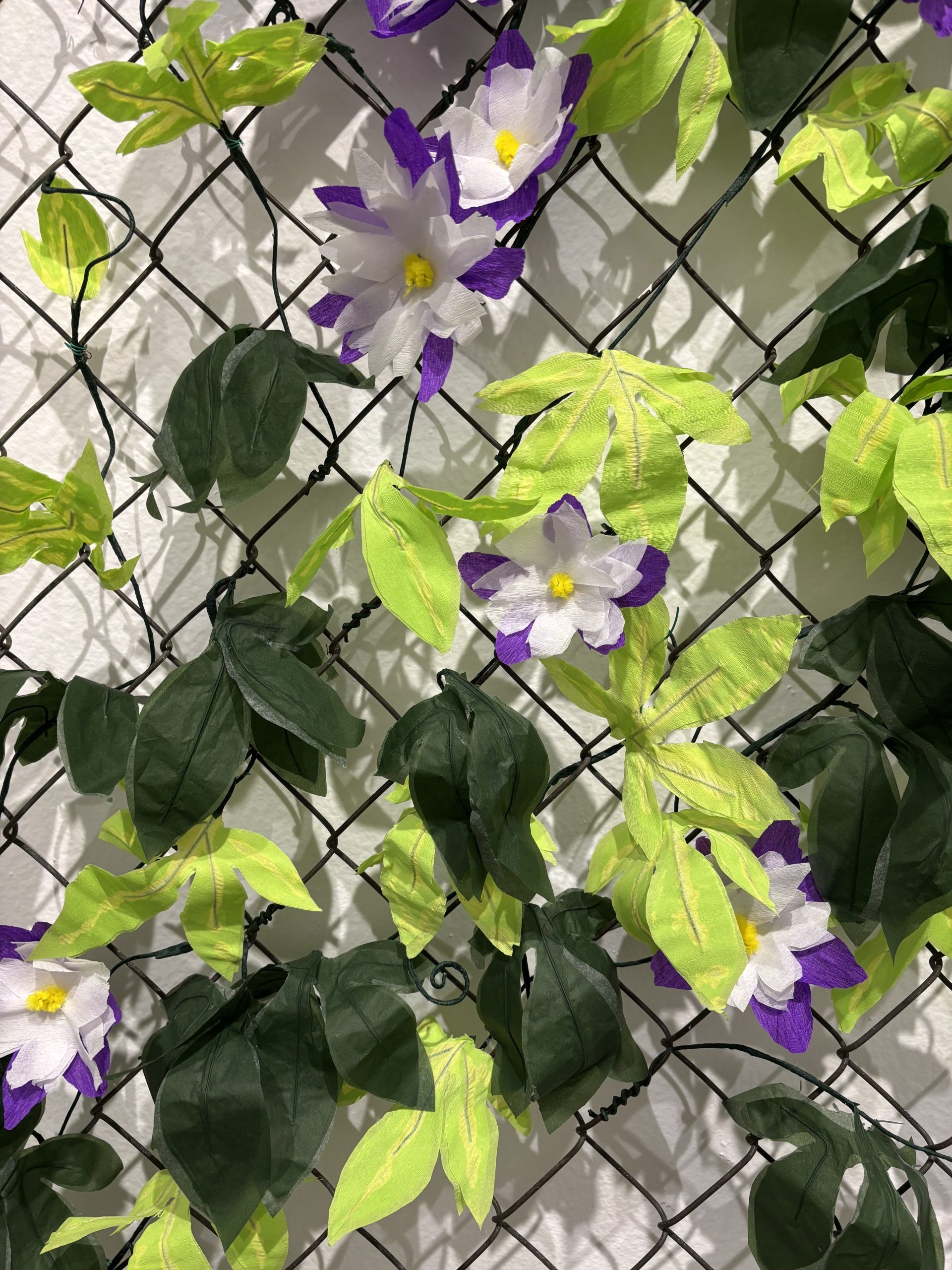 Paper passionflowers in purple and white with yellow centers, alongside crepe paper leaves, arranged on a chain link fence.