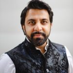 Sonny K. Mehta, a Houston-based Qawwali musician and community documentarian, photographed in a headshot. He has dark hair, a full beard, and wears a patterned black vest over a white collared shirt against a light gray background.