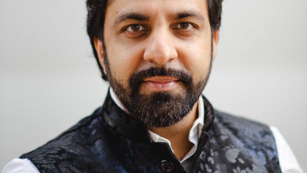 Sonny K. Mehta, a Houston-based Qawwali musician and community documentarian, photographed in a headshot. He has dark hair, a full beard, and wears a patterned black vest over a white collared shirt against a light gray background.