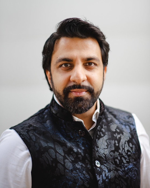 Sonny K. Mehta, a Houston-based Qawwali musician and community documentarian, photographed in a headshot. He has dark hair, a full beard, and wears a patterned black vest over a white collared shirt against a light gray background.