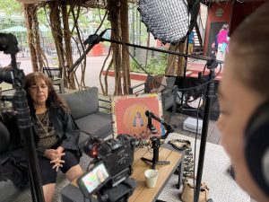 image0 Mary Jane Garza speaks during a filmed interview on a shaded outdoor patio. A microphone boom, camera, and softbox light surround her, and a devotional painting of a robed figure holding a child rests on the table beside her.