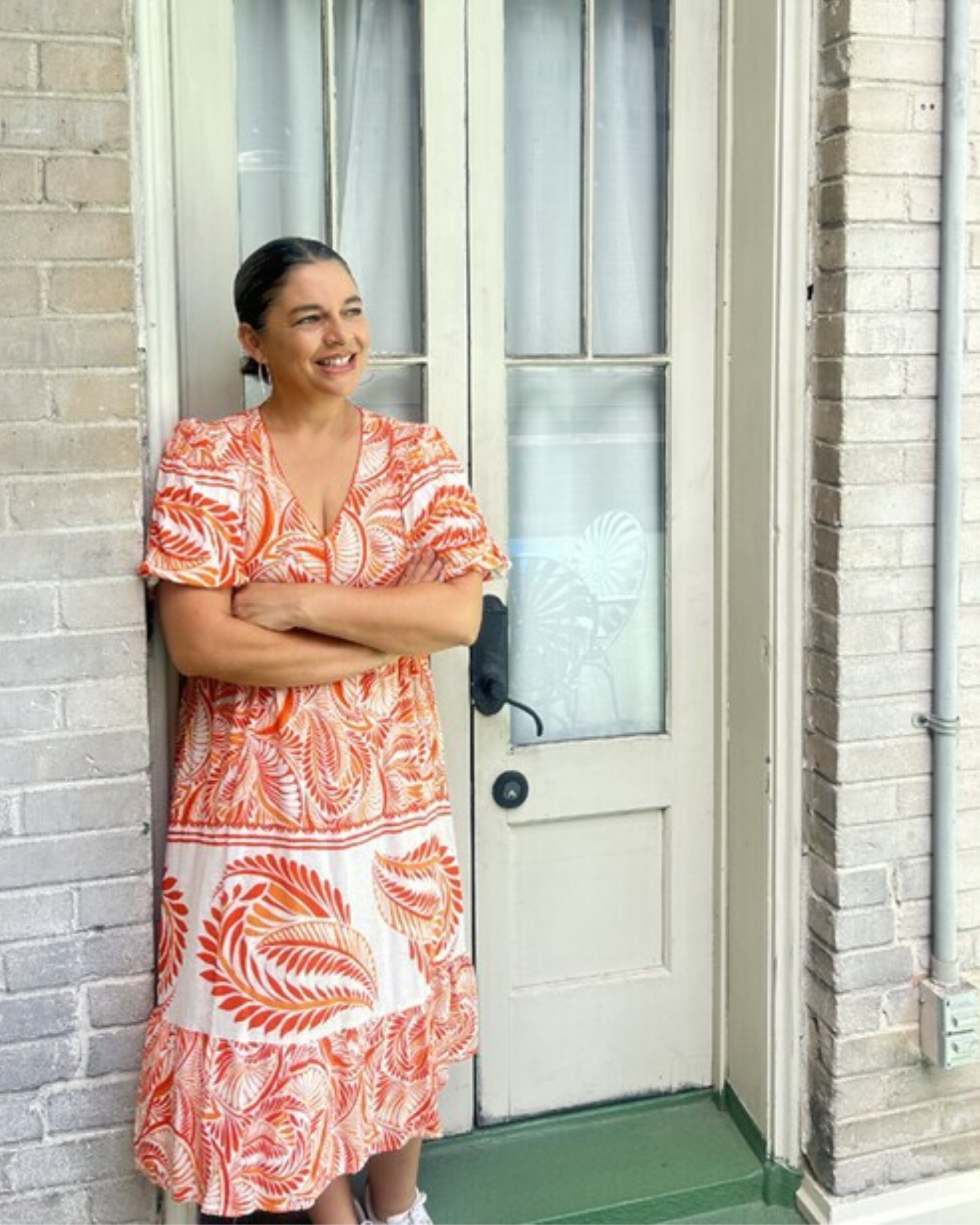 Oddett Garza Oddett Garza stands in a doorway with her arms crossed, smiling and looking off to the side. She wears an orange and white patterned dress with short sleeves, and her dark hair is pulled back. Behind her is a white-painted wooden door with glass panes, set into a light brick wall with a green painted step.