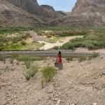 A person with long dark hair, wearing a red top, red backpack, and a long brown tiered skirt with red trim, walks down a rocky desert slope toward a two-lane road. Beyond the road, the Río Grande curves through green brush, with steep canyon mountains rising in the background under a pale sky.