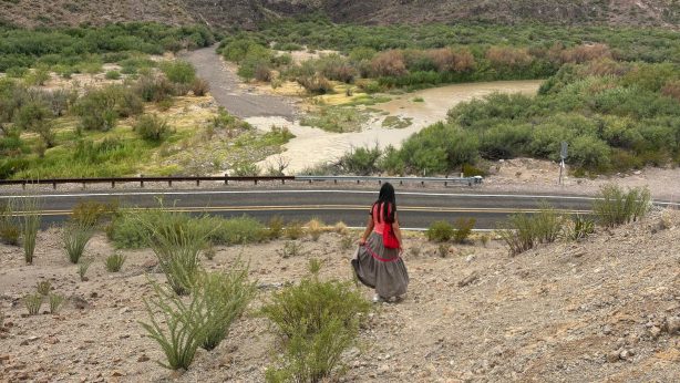 A person with long dark hair, wearing a red top, red backpack, and a long brown tiered skirt with red trim, walks down a rocky desert slope toward a two-lane road. Beyond the road, the Río Grande curves through green brush, with steep canyon mountains rising in the background under a pale sky.