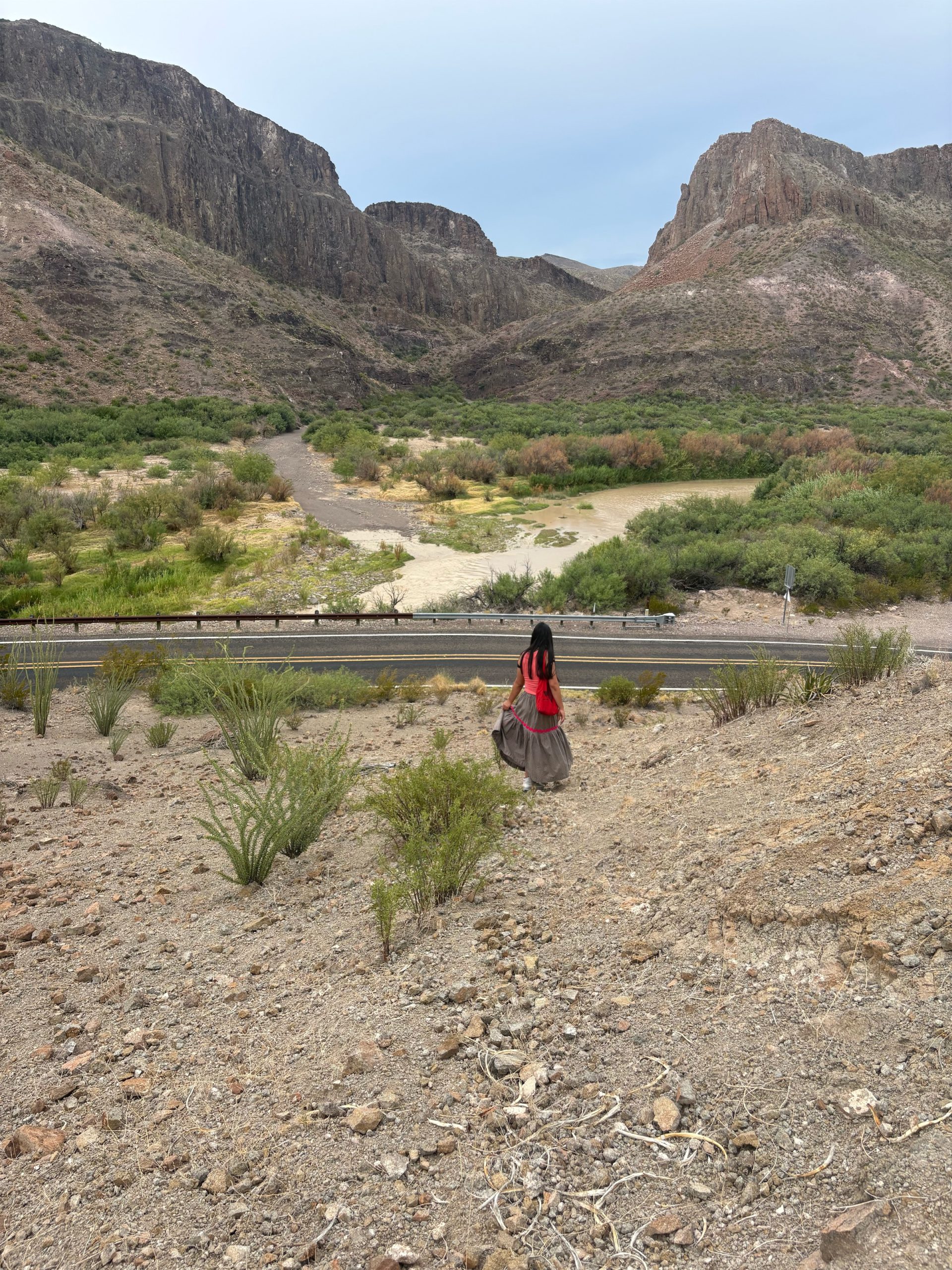 A person with long dark hair, wearing a red top, red backpack, and a long brown tiered skirt with red trim, walks down a rocky desert slope toward a two-lane road. Beyond the road, the Río Grande curves through green brush, with steep canyon mountains rising in the background under a pale sky.