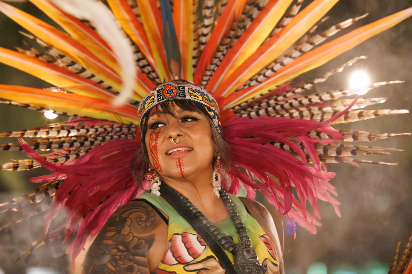 [Laura Yohualtlahuiz] dances with one arm extended and a wide smile, wearing yellow and red face paint, a sheer black feathered top, and several strands of jade beaded necklaces with carved Mesoamerican pendants. A woven basket rests on one arm. Colorful papel picado banners and string lights fill the background. Photo by Mel Cardon.