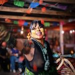 [Laura Yohualtlahuiz] wears an elaborate Mexica danza headdress with long feathers radiating outward in orange, red, and pink alongside brown-and-white patterned feathers. A beaded headband with colorful floral and geometric designs frames her face, which is marked with red linear face paint. She wears a gold septum ring and shell earrings, with a tattoo visible on one arm and a green and yellow patterned top. Warm lights glow in the background.