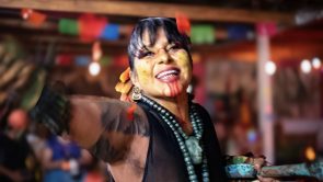 [Laura Yohualtlahuiz] wears an elaborate Mexica danza headdress with long feathers radiating outward in orange, red, and pink alongside brown-and-white patterned feathers. A beaded headband with colorful floral and geometric designs frames her face, which is marked with red linear face paint. She wears a gold septum ring and shell earrings, with a tattoo visible on one arm and a green and yellow patterned top. Warm lights glow in the background.