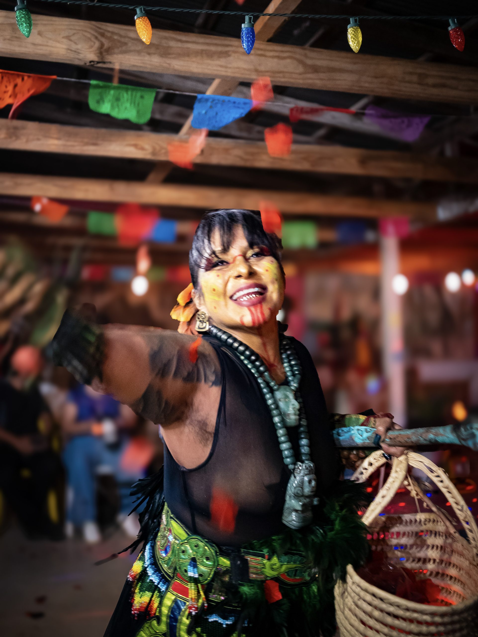 [Laura Yohualtlahuiz] wears an elaborate Mexica danza headdress with long feathers radiating outward in orange, red, and pink alongside brown-and-white patterned feathers. A beaded headband with colorful floral and geometric designs frames her face, which is marked with red linear face paint. She wears a gold septum ring and shell earrings, with a tattoo visible on one arm and a green and yellow patterned top. Warm lights glow in the background.